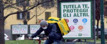 A food delivery man wearin a  protective mask rides his bike past posters reading in italian “Protect yourself, protect others” , Milan, Italy, 25 March 2020.The World Health Organization (WHO) has said that the total figures of those infected by the pandemic COVID-19 disease caused by the SARS-CoV-2 coronavirus throughout Italy may soon see their peak once the effects of the strict national lockdown implemented on 09 March start to kick in. ANSA/DANIEL DAL ZENNARO